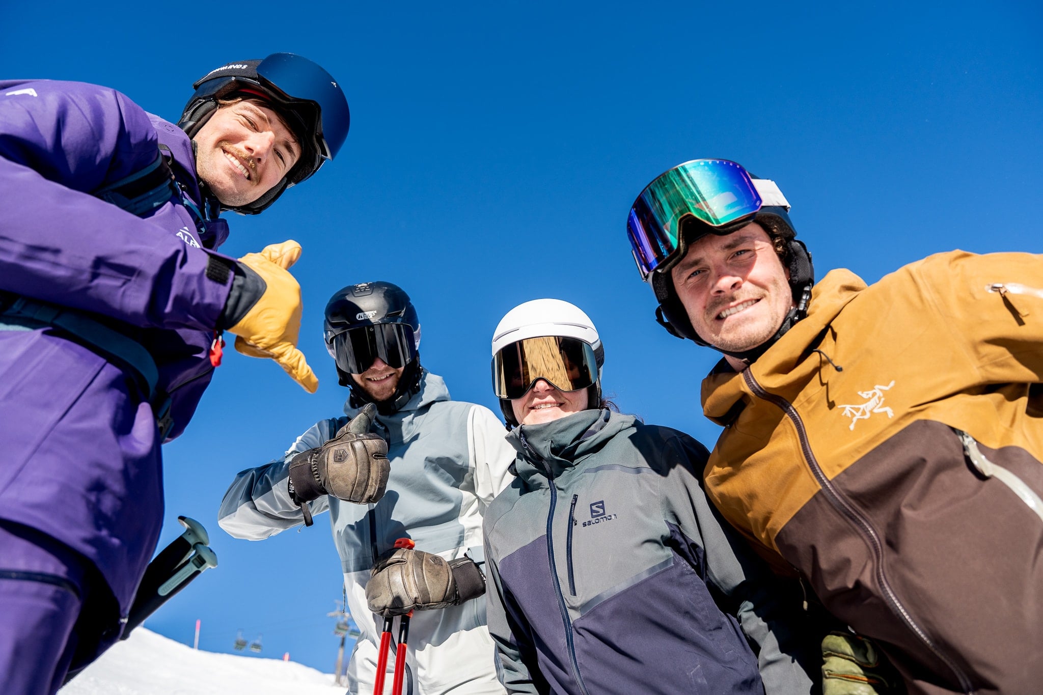 Vier Skifahrende, eine davon ein flow Guide in lila Albona-Jacke, lächeln vor tiefblauem Arlberg-Himmel in die Kamera.
