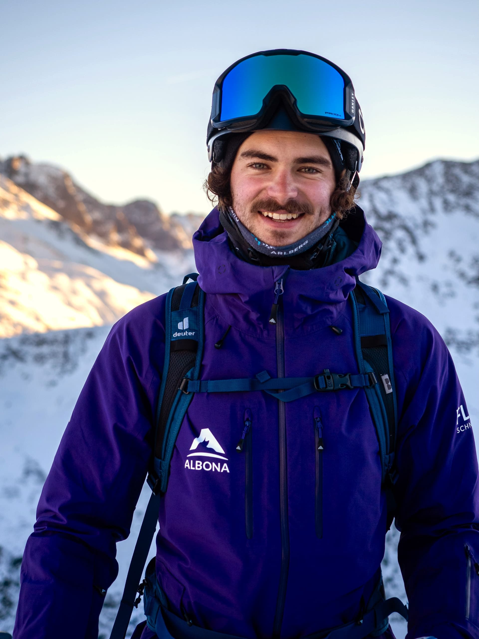 A flow guide in a purple Albona jacket and blue mirrored goggles smiles broadly, the sunlit Arlberg peaks behind him.