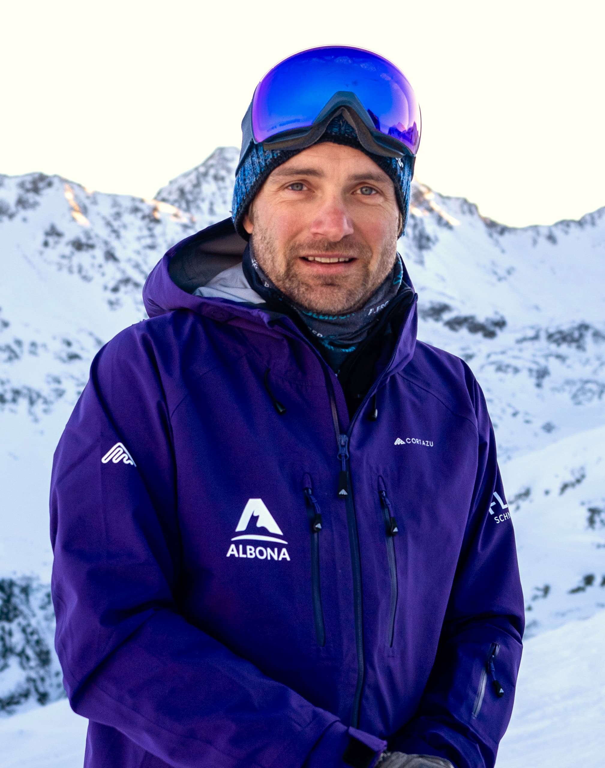 A flow guide in a purple Albona jacket, beanie and blue mirrored goggles, smiles in front of the snowy Arlberg mountains.