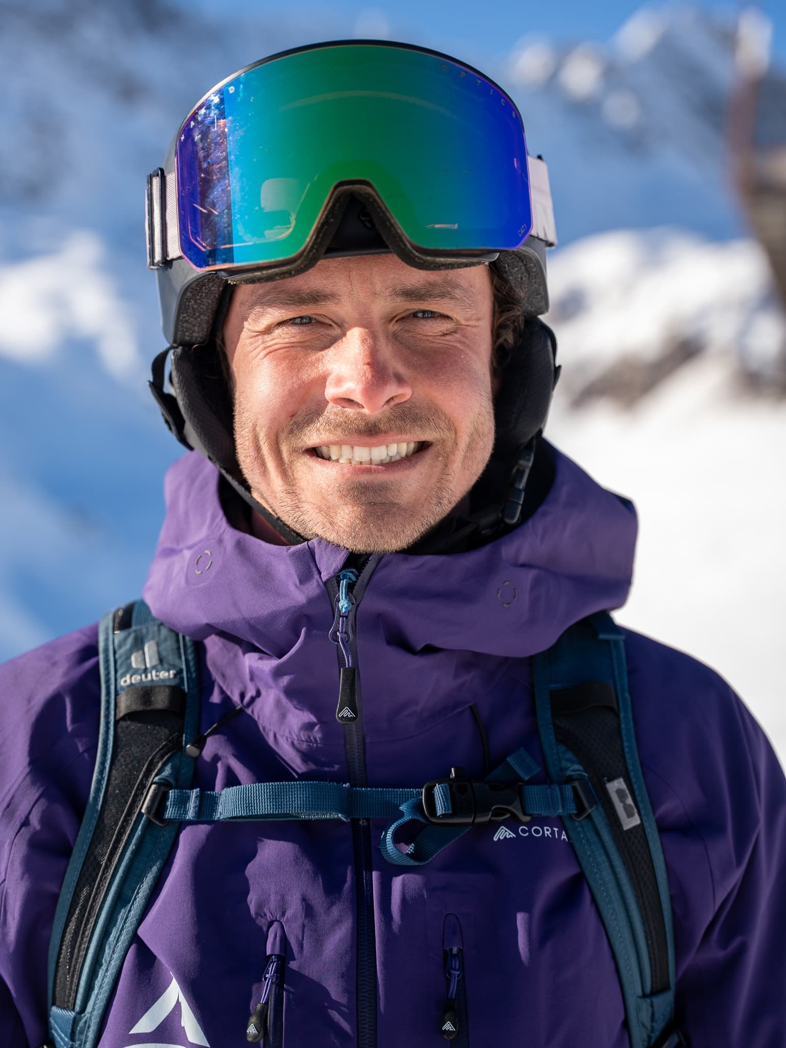 A flow guide in a purple Albona jacket with green mirrored goggles on his helmet, a snowy Arlberg face behind him.