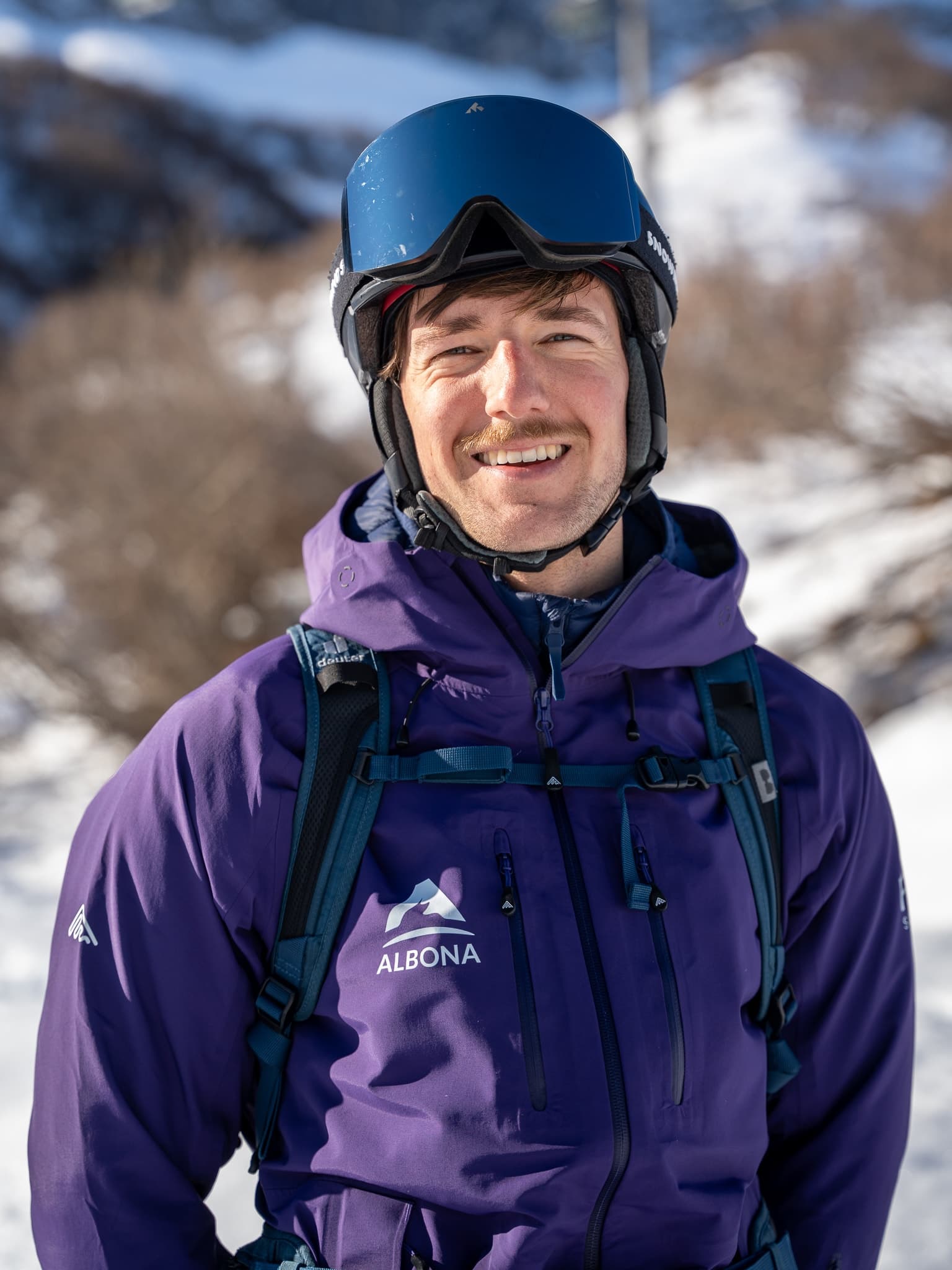 A flow guide in a purple Albona jacket, goggles pushed up on his blue helmet, smiles into the camera above Stuben am Arlberg.